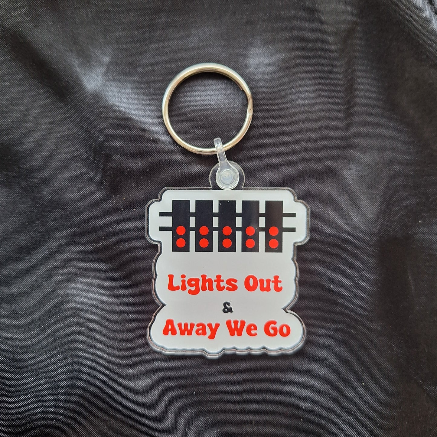 An acrylic keyring with a featuring a white and red design with text 'LIGHTS OUT & AWAY WE GO' on a white background.