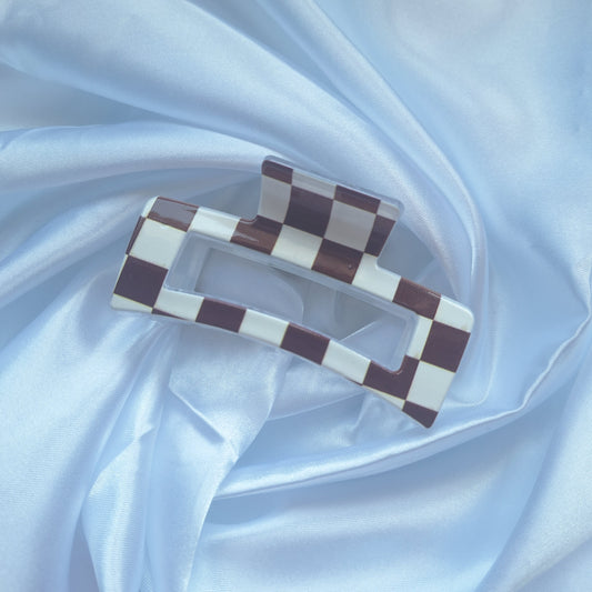 Brown and white checkered acrylic hair claw clip inspired by racing flags displayed on white satin fabric.